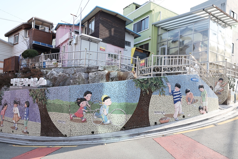 Foto di atas menunjukkan sisi depan Desa Batu Nisan Ami-dong yang terletak di Seo-gu, Kota Busan. Desa ini dibangun oleh para pengungsi yang tiba di Kota Busan dengan menggunakan batu-batu nisan yang tersisa karena lahan desa tersebut awalnya digunakan sebagai pemakaman umum warga Jepang sejak tahun 1906.  