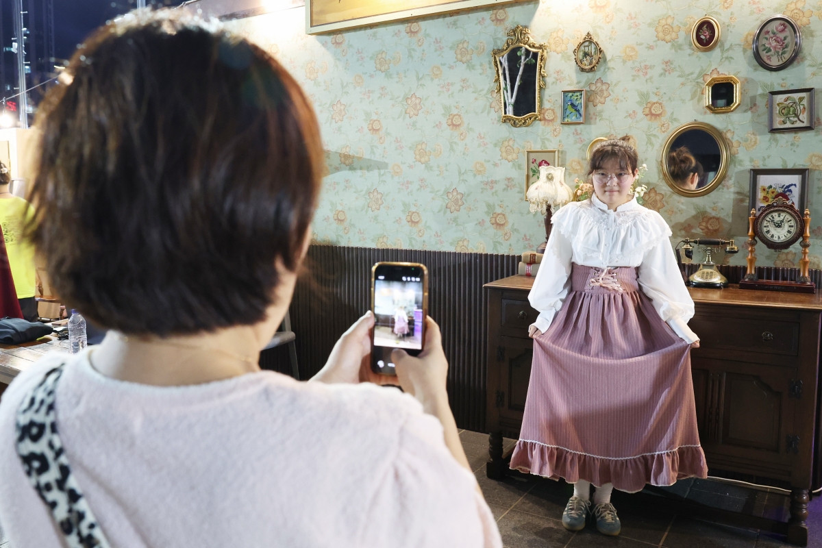 Seorang ibu memotret anaknya yang mengenakan baju dengan model tahun 1950-an dalam Busan Wartime Capital Heritage Night yang digelar pada tanggal 25 Juli 2026 di Alun-alun Yurari, Jung-gu, Busan. Zona foto dipasang di berbagai sudut alun-alun untuk membuat pengunjung merasakan suasana Busan pada tahun 1950-an. 