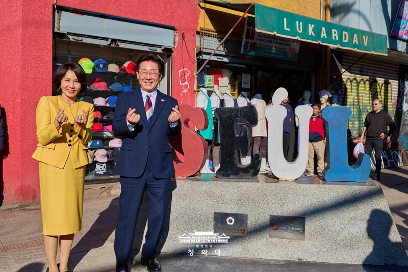 Presiden Lee Jae Myung dan Ibu Negara Kim Hea Kyung memberikan simbol hati dengan jari tangan mereka di depan instalasi Jalan Seoul pada tanggal 30 Juli 2026 (waktu setempat) di Distrik Patronato, Santiago, Cili. Presiden dan Ibu Negara mengunjungi serta berkomunikasi dengan diaspora Korea di Cili melalui kunjungan langsung ke Distrik Patronato dengan banyak toko yang dimiliki oleh diaspora Korea.