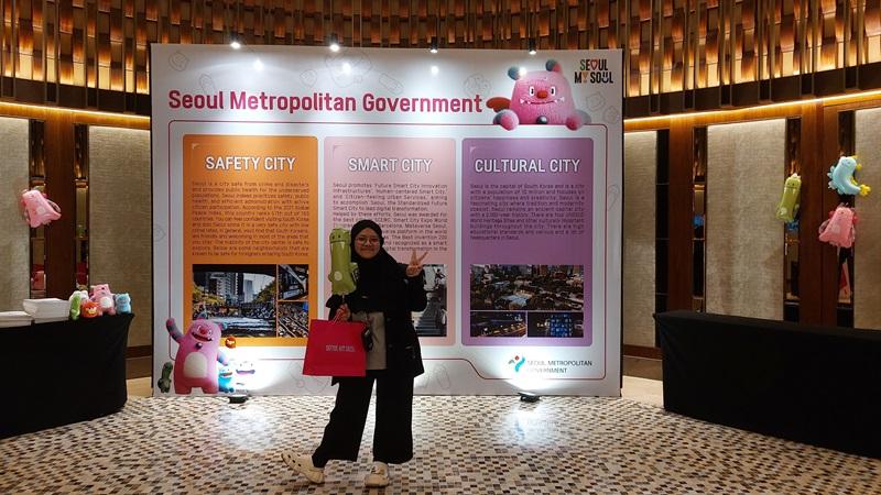 Penulis menghadiri pameran pendidikan Study in Seoul dan berfoto di depan papan informasi mengenai Kota Metropolitan Seoul sembari membawa tas kertas berisi suvenir acara pameran. 