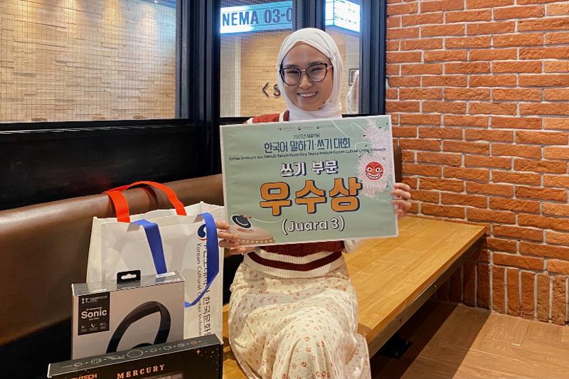 Potret penulis memegang papan penghargaan bertuliskan Juara 3 dari Lomba Menulis dalam Bahasa Korea.  