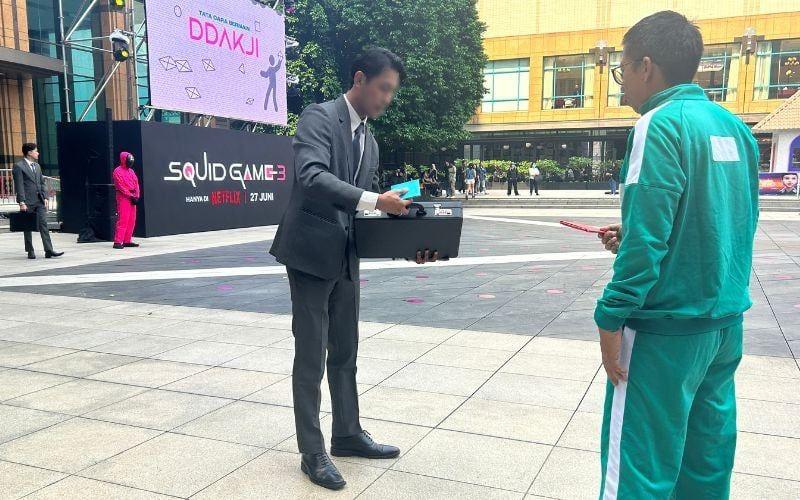 Pengunjung bermain ddakji bersama petugas berkostum perekrut seperti dalam adegan pembuka serial Squid Game. 