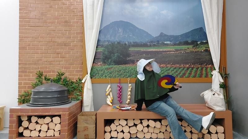Penulis berfoto di instalasi dapur tradisional Korea yang merepresentasikan suasana musim panas di desa, lengkap dengan tungku, bangku kayu, dan latar pemandangan sawah. (Maulia Resta Mardaningtias) 