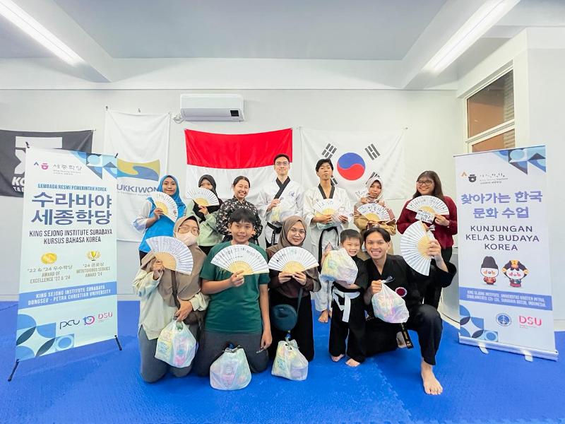 Seluruh peserta dan guru berfoto bersama dengan kipas bertuliskan hangeul dalam Kunjungan Kelas Budaya Korea yang digelar pada tanggal 20 Mei 2026 di Akademi HKAA, Surabaya. (Humas HKAA) 